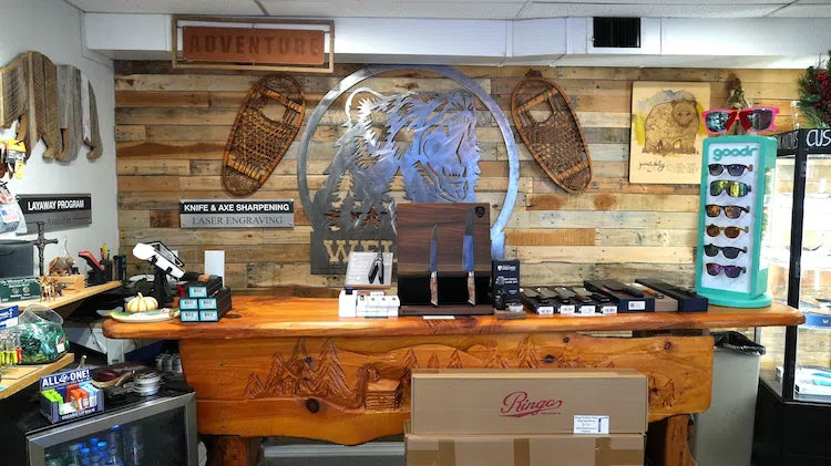 A rustic wooden counter in a shop interior with a large metal Native American profile sculpture mounted on a wood-plank wall. Snowshoes hang on the wall, framed art is visible, and a turquoise eyeglasses display stand stands to the right.