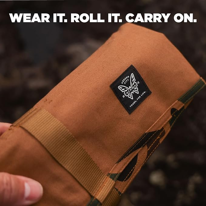 A close-up of a brown canvas roll-up pouch or sleeve with a black brand patch reading 'Proudly Carry On' and a thumb holding it. Large white text at the top says 'WEAR IT.