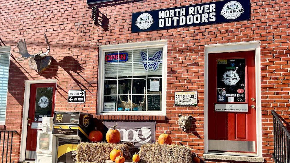 North River Outdoors storefront on a red brick building, with a large black sign above the window, a red door, an open neon sign, hay bales with pumpkins, and outdoor decor.