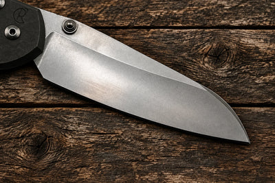 A close-up of a folding knife blade with a satin finish, showing a black handle scale with a circular logo, set against a plain light gray background.