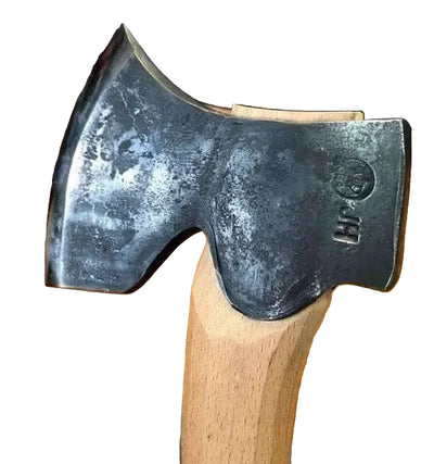 A close-up of a hand axe or hatchet head with a dark forged steel blade and a light natural wood handle, resting on a wooden surface.